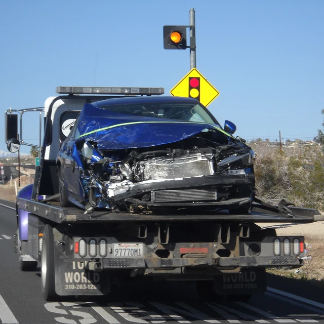 A crashed blue car on the back of a tow truck under a clear sky.